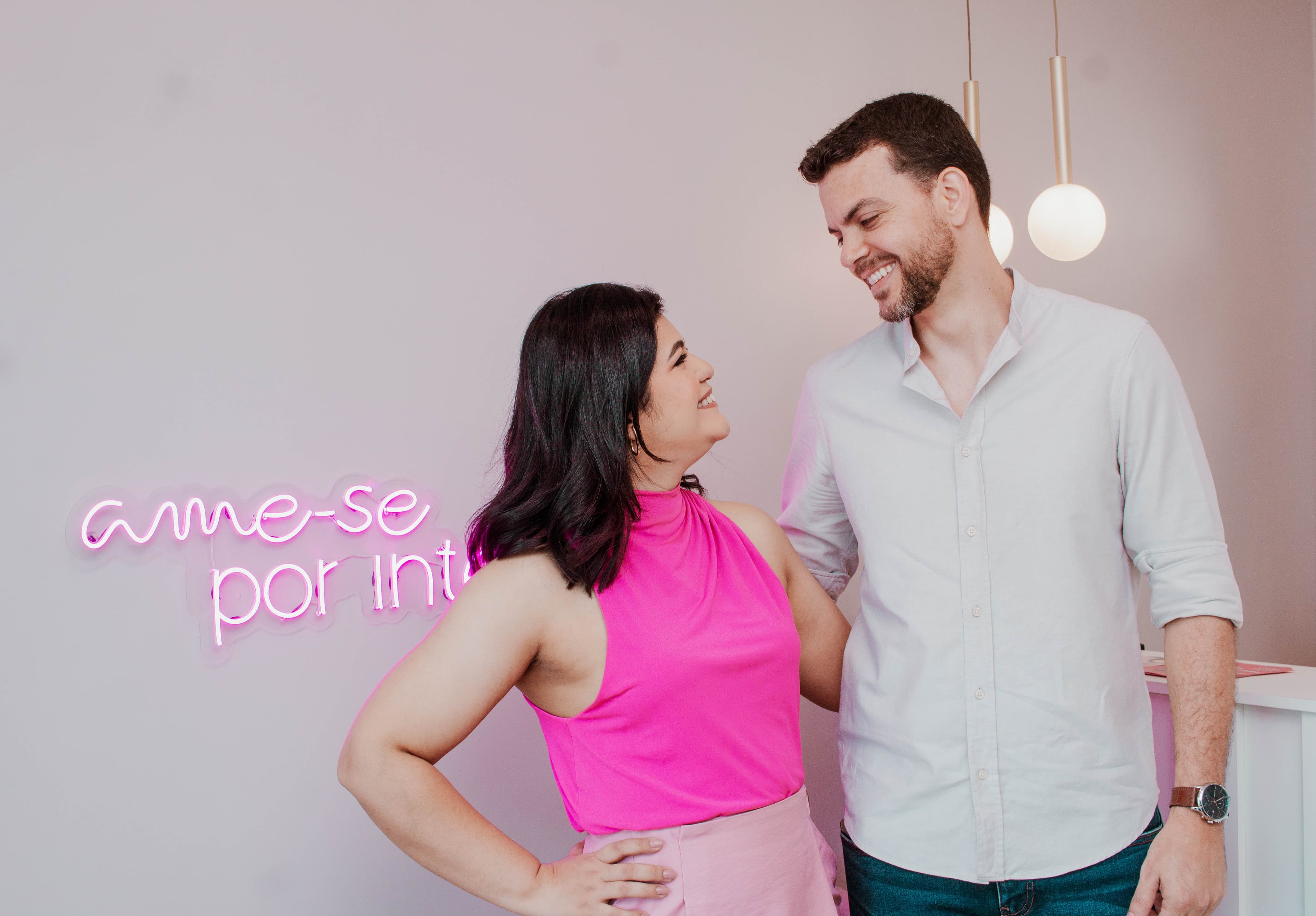 Casal sorridente conversando em frente ao letreiro em neon rosa 'ame-se por inteiro' em uma unidade Clube Depil